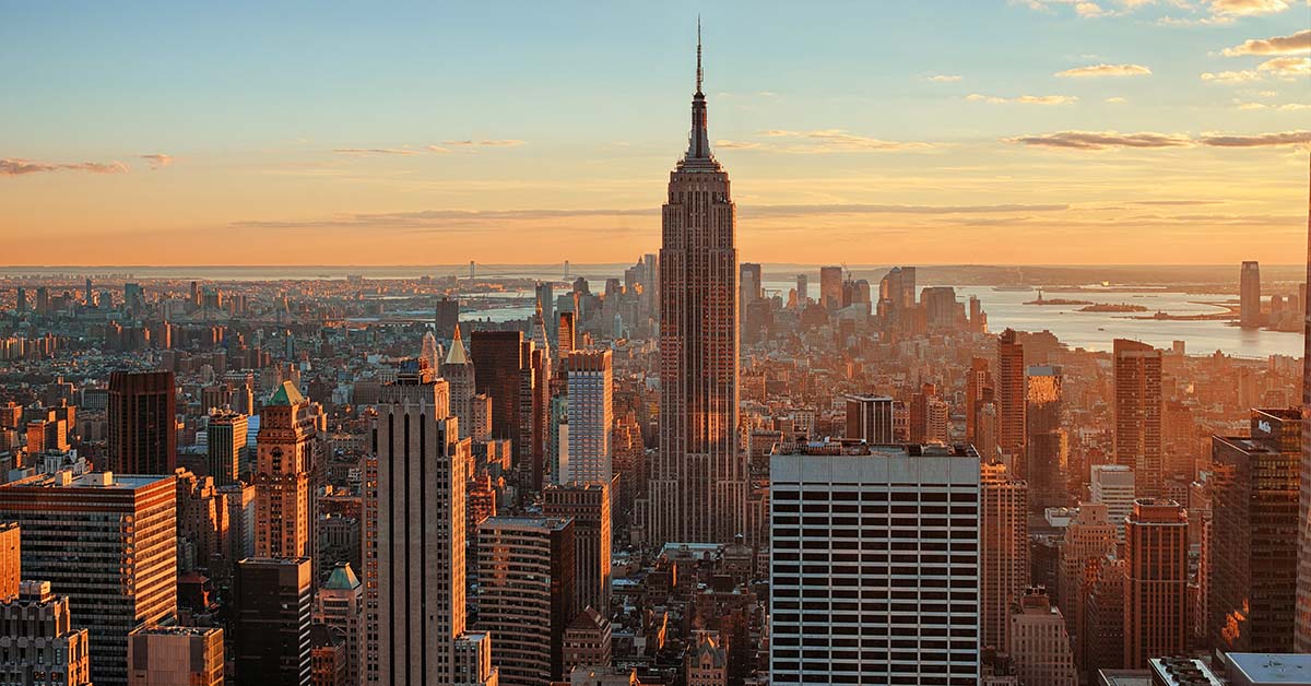 Aerial view of the Manhattan skyline at sunset, with the Empire State Building rising at the center above dense skyscrapers, bathed in warm golden light with the harbor visible in the distance.