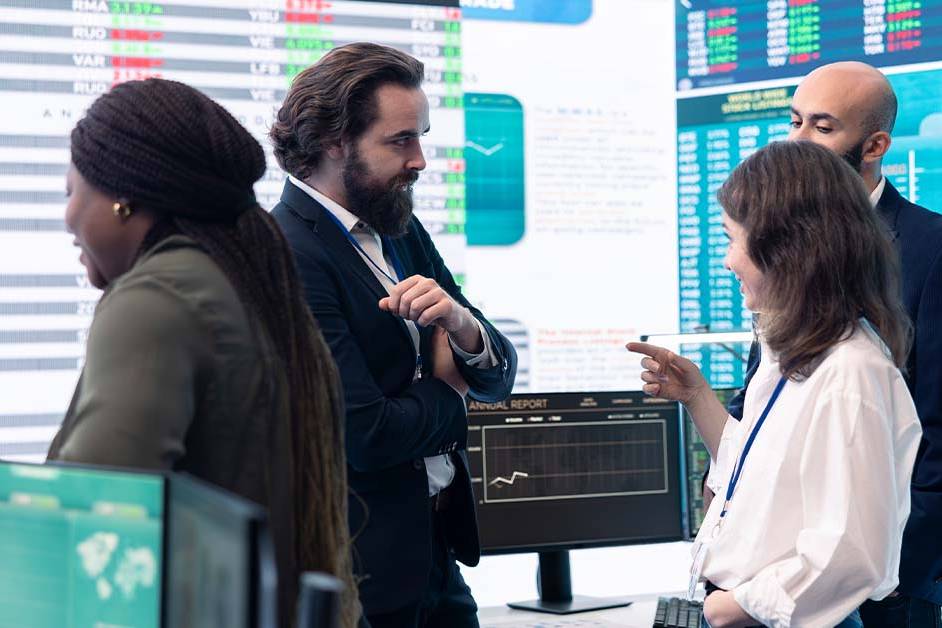 A diverse team of four financial executives discussing data in front of a wall of digital market charts, featured image for the article Therapy for Bay Area IPO-Tier CFOs.