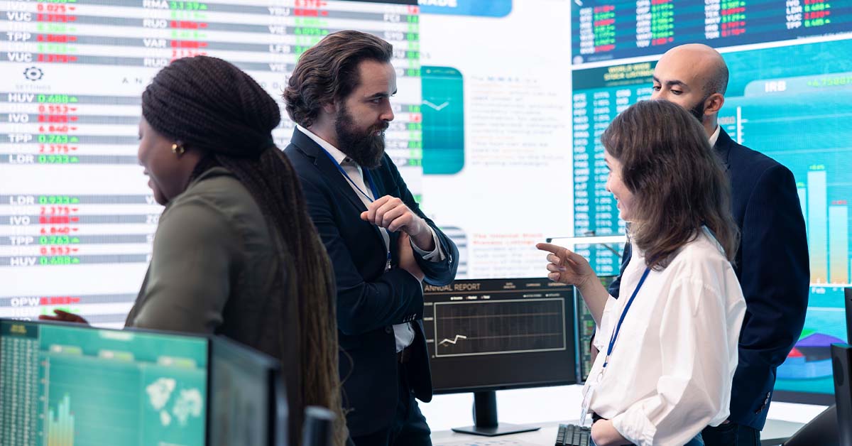 A diverse team of four financial executives discussing data in front of a wall of digital market charts, featured image for the article Therapy for Bay Area IPO-Tier CFOs.