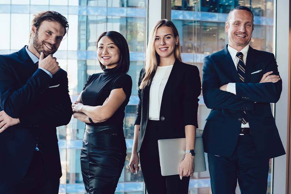 Four confident, well-dressed business professionals in dark suits stand together smiling in front of floor-to-ceiling windows overlooking a city skyline in a high-rise office.