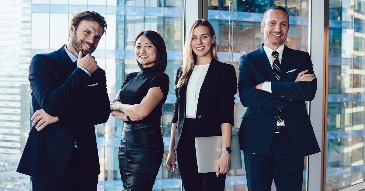 Four confident, well-dressed business professionals in dark suits stand together smiling in front of floor-to-ceiling windows overlooking a city skyline in a high-rise office.