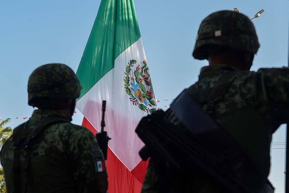 Two armed soldiers in camouflage standing in front of a large Mexican flag, featured image for the article Therapy for Border Patrol Agents.