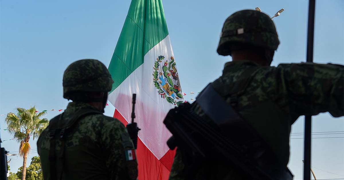 Two armed soldiers in camouflage standing in front of a large Mexican flag, featured image for the article Therapy for Border Patrol Agents.