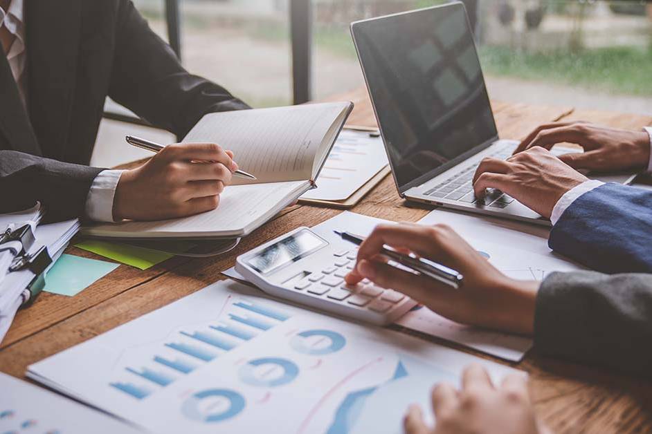 Three professionals reviewing financial charts, using a calculator, and typing on a laptop at a wooden desk, featured image for the article Therapy for CPAs With Board Reporting Concerns.