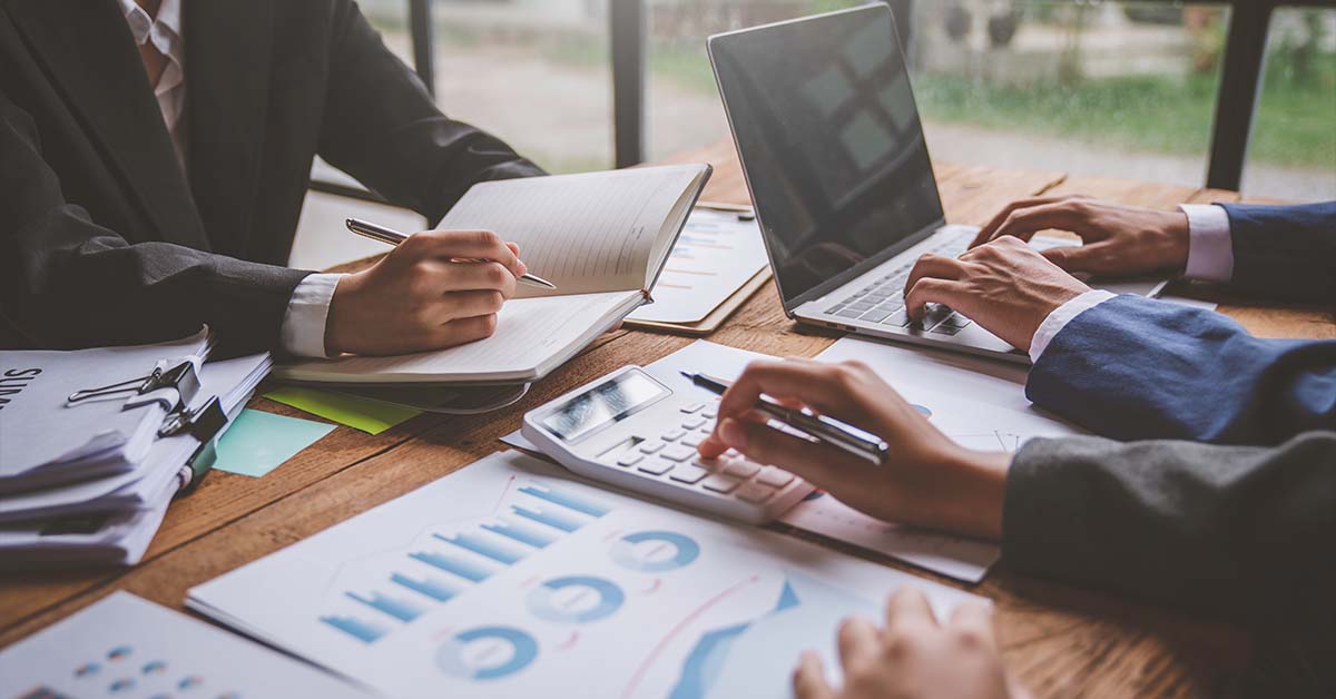 Three professionals reviewing financial charts, using a calculator, and typing on a laptop at a wooden desk, featured image for the article Therapy for CPAs With Board Reporting Concerns.