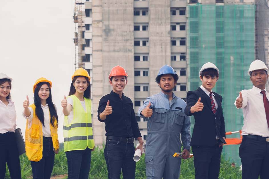 A diverse group of seven construction and business professionals in hard hats, safety vests, and suits give a thumbs up in front of a high-rise building under construction.