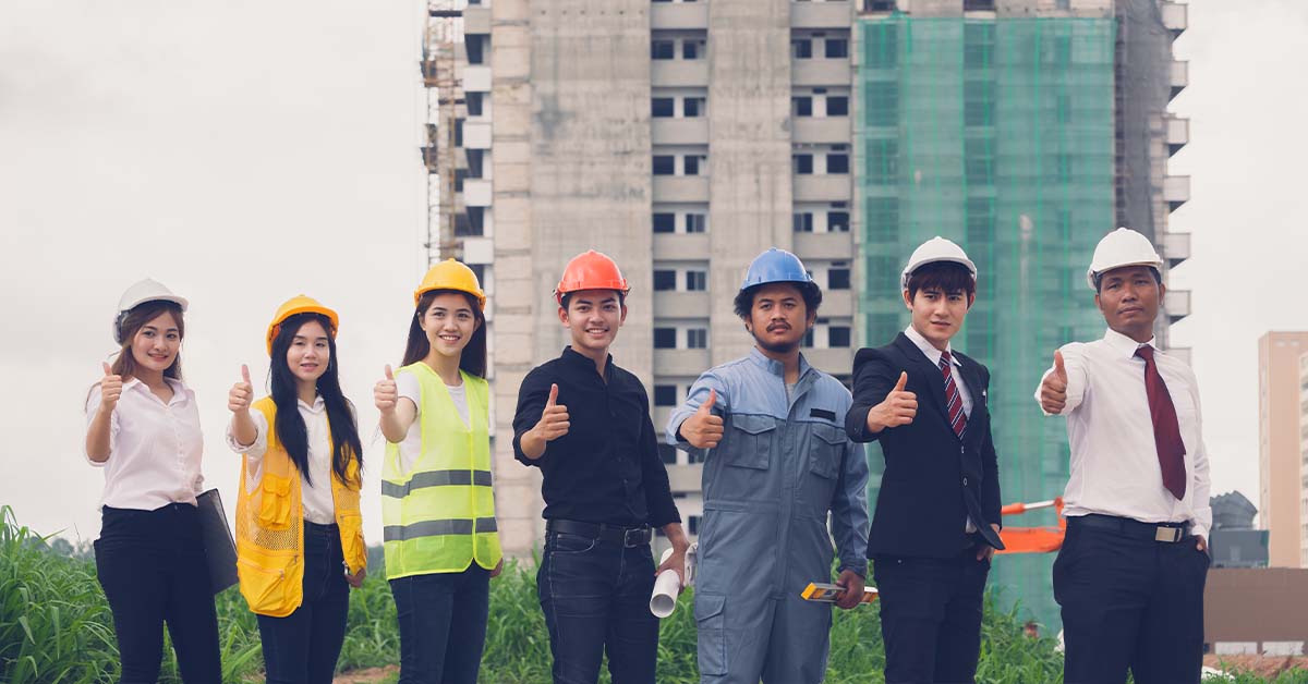 A diverse group of seven construction and business professionals in hard hats, safety vests, and suits give a thumbs up in front of a high-rise building under construction.
