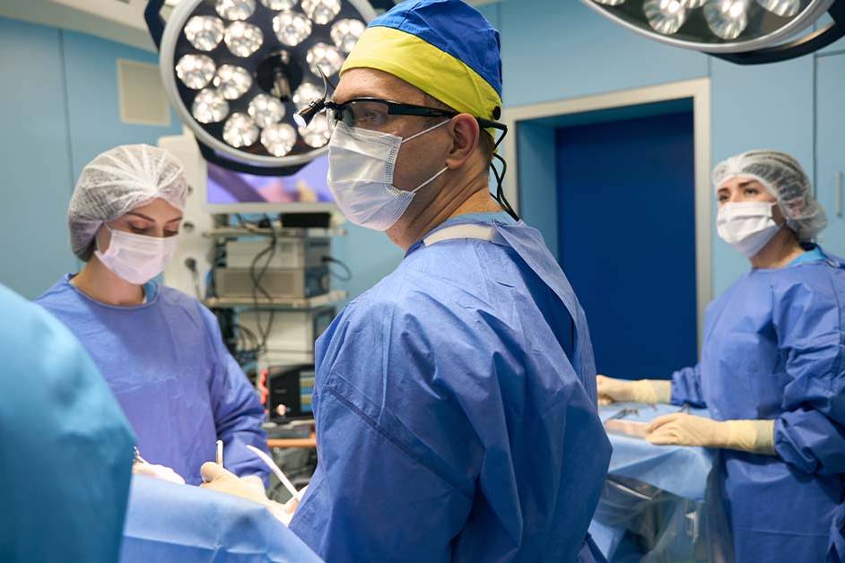 A surgeon in blue scrubs, surgical cap, mask, and magnifying loupes stands in an operating room with two gowned team members under bright surgical lights during a procedure.