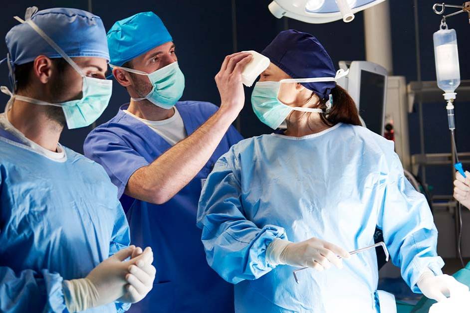 An assistant wiping the brow of a female surgeon in the middle of a tense operation, featured image for the article Therapy for Chicago Surgeons.