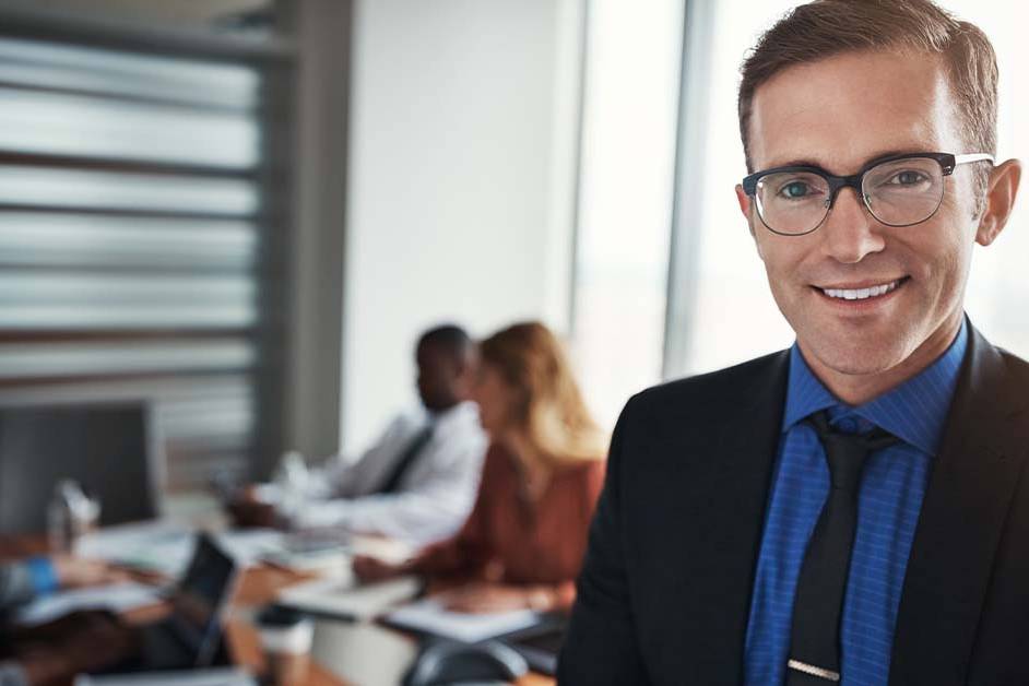 A smiling male executive in a suit and glasses standing in front of a blurred boardroom meeting, featured image for the article Therapy for Chief Compliance Officers in Financial Services.