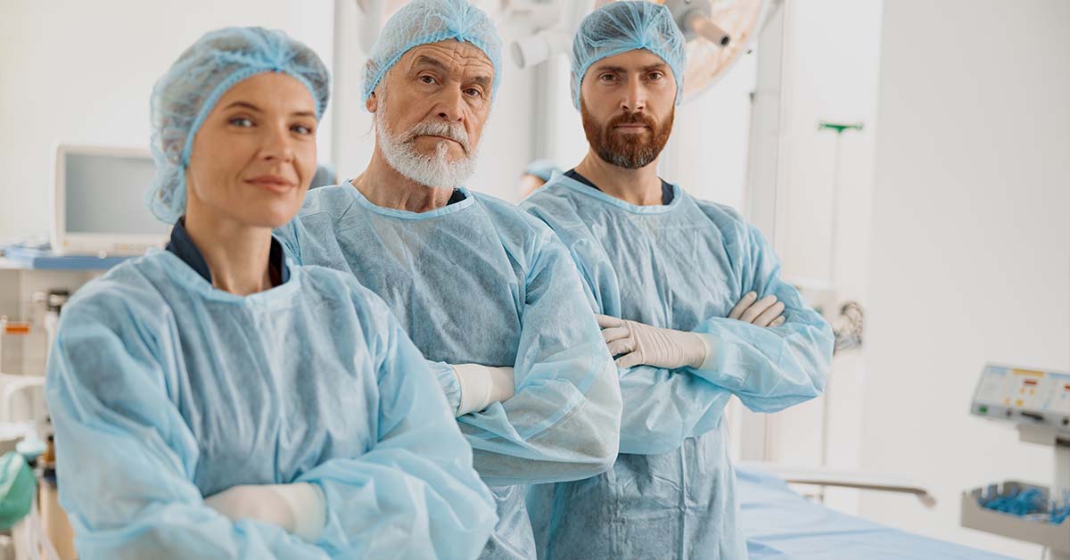 Three medical professionals in blue surgical gowns and caps standing with arms crossed in a clinical setting, featured image for the article Therapy for Chronic Insomnia in Surgeons and ICU Physicians.