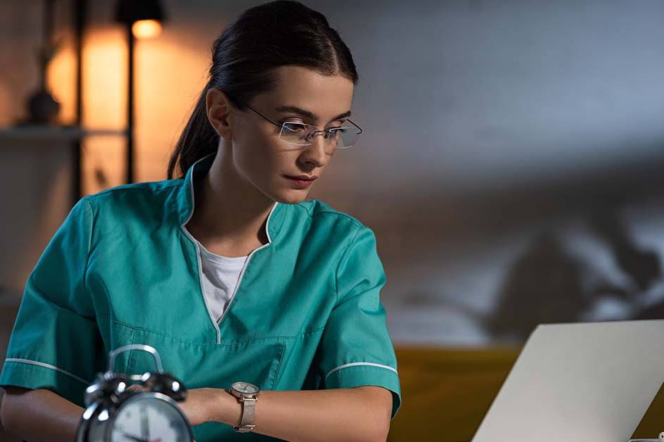 A focused female physician in green scrubs working on a laptop late at night with an alarm clock in the foreground, featured image for the article Therapy for Circadian Disruption in Shift-Working Physicians.