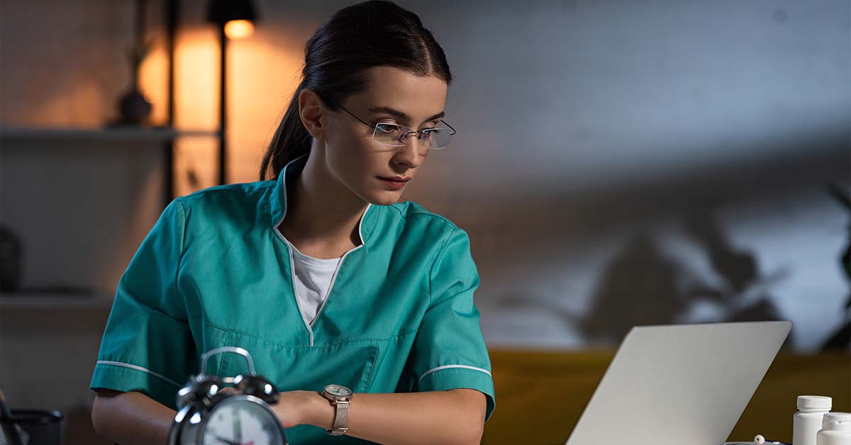 A focused female physician in green scrubs working on a laptop late at night with an alarm clock in the foreground, featured image for the article Therapy for Circadian Disruption in Shift-Working Physicians.