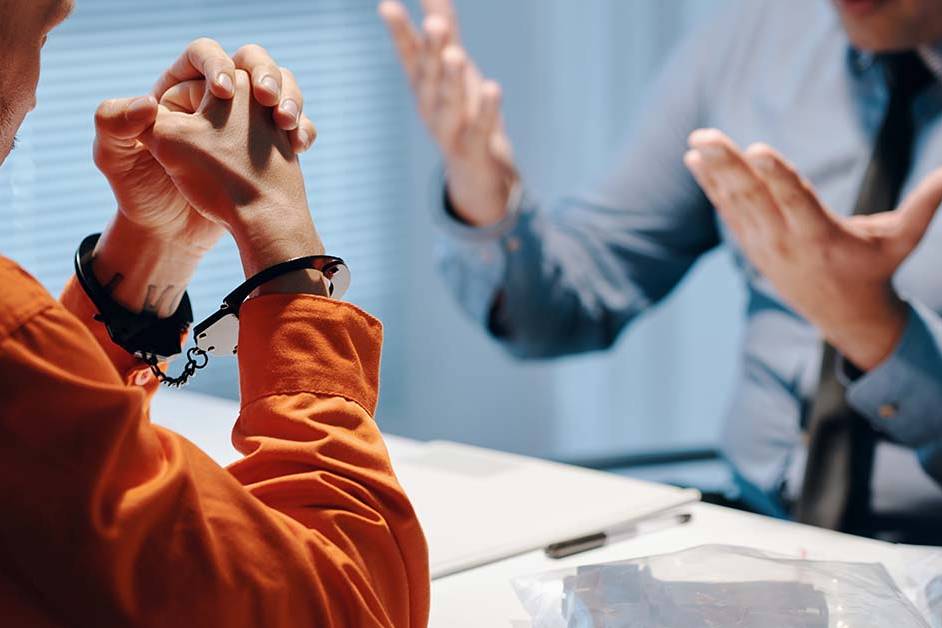 A handcuffed man in an orange jumpsuit sitting across from an attorney gesturing at a white table, featured image for the article Therapy for Criminal Defense Attorneys.