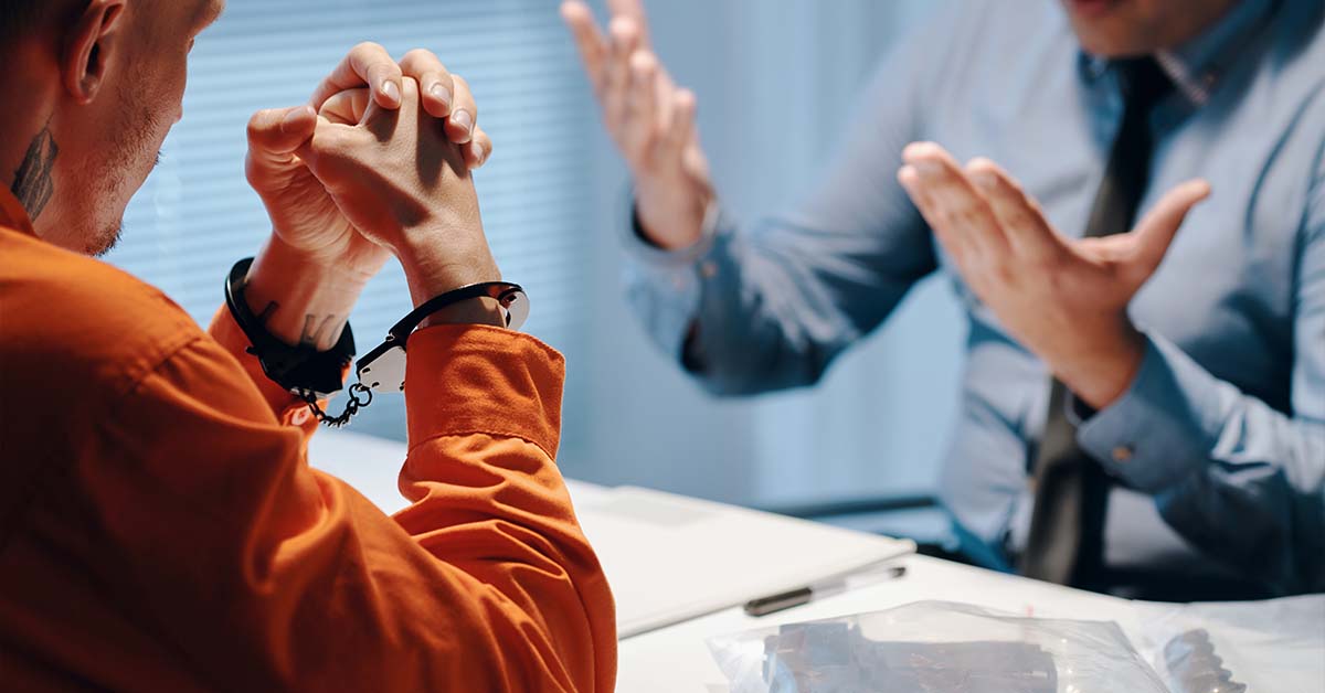 A handcuffed man in an orange jumpsuit sitting across from an attorney gesturing at a white table, featured image for the article Therapy for Criminal Defense Attorneys.