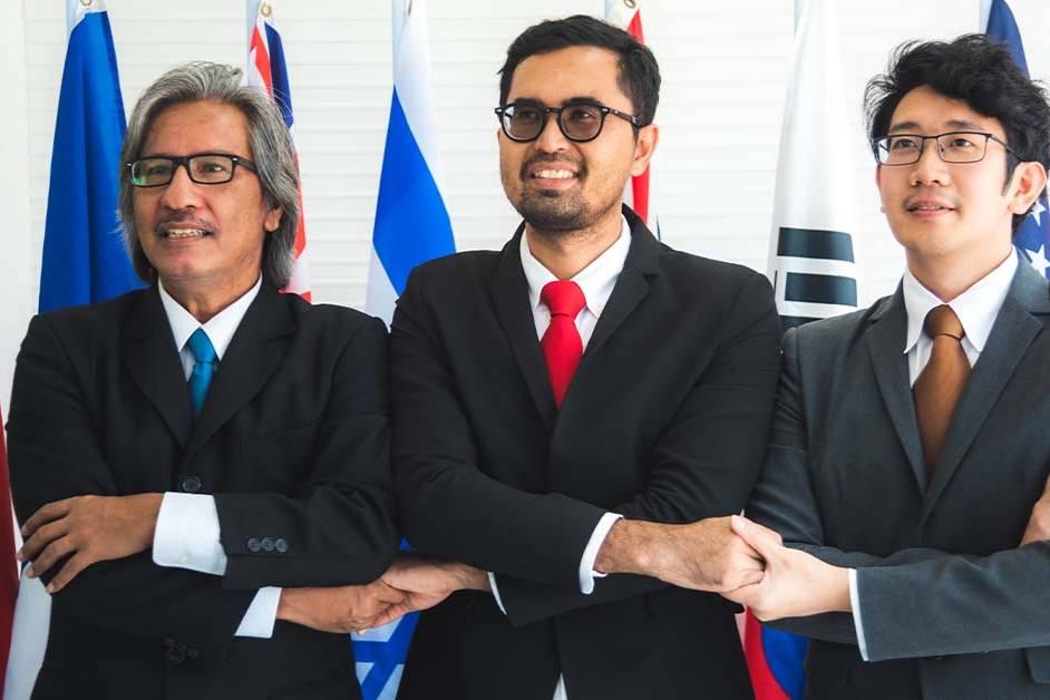 Three diverse political officials in suits performing a cross-armed handshake in front of international flags, featured image for the article Therapy for DC Diplomats Between Posts.