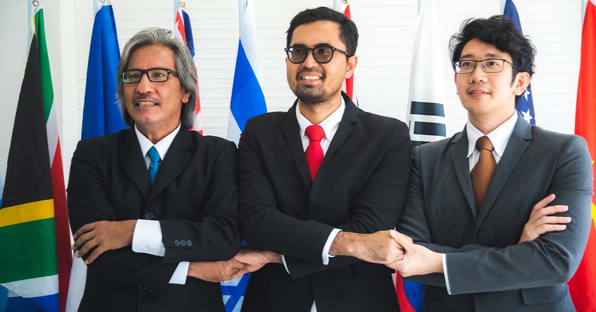 Three diverse political officials in suits performing a cross-armed handshake in front of international flags, featured image for the article Therapy for DC Diplomats Between Posts.