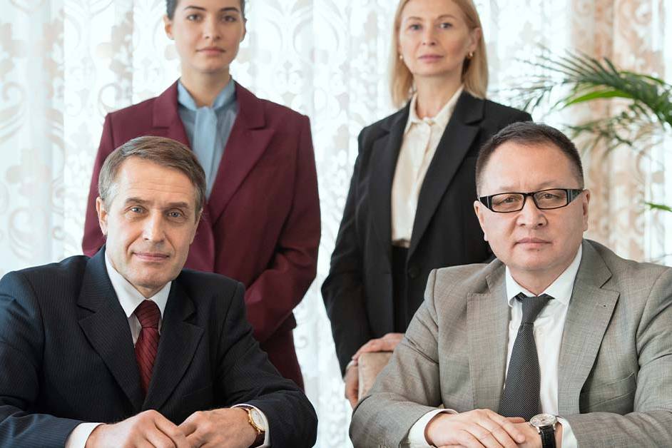 Four serious business professionals in formal suits pose in a formal portrait, two seated men in front and two women standing behind, in an elegant room with patterned drapes.