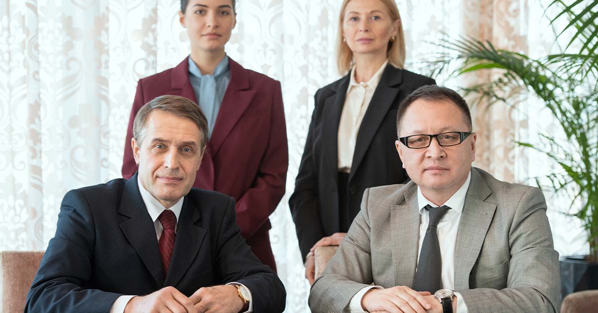 Four serious business professionals in formal suits pose in a formal portrait, two seated men in front and two women standing behind, in an elegant room with patterned drapes.