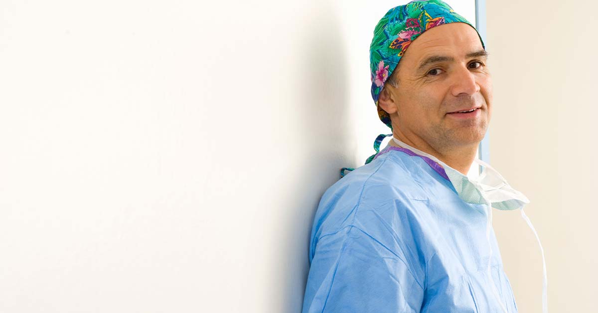 A middle-aged male surgeon in blue scrubs and a colorful floral surgical cap leans against a white wall with his mask lowered, smiling faintly during a quiet moment, with ample white copy space.