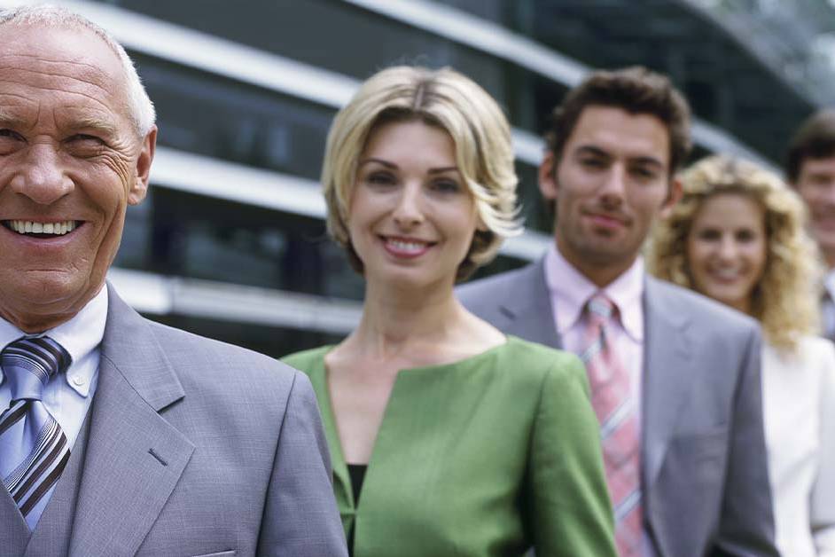 A smiling senior male executive in a grey suit standing in front of a line of blurred colleagues outside a glass building, featured image for the article Therapy for Executives Between Roles.