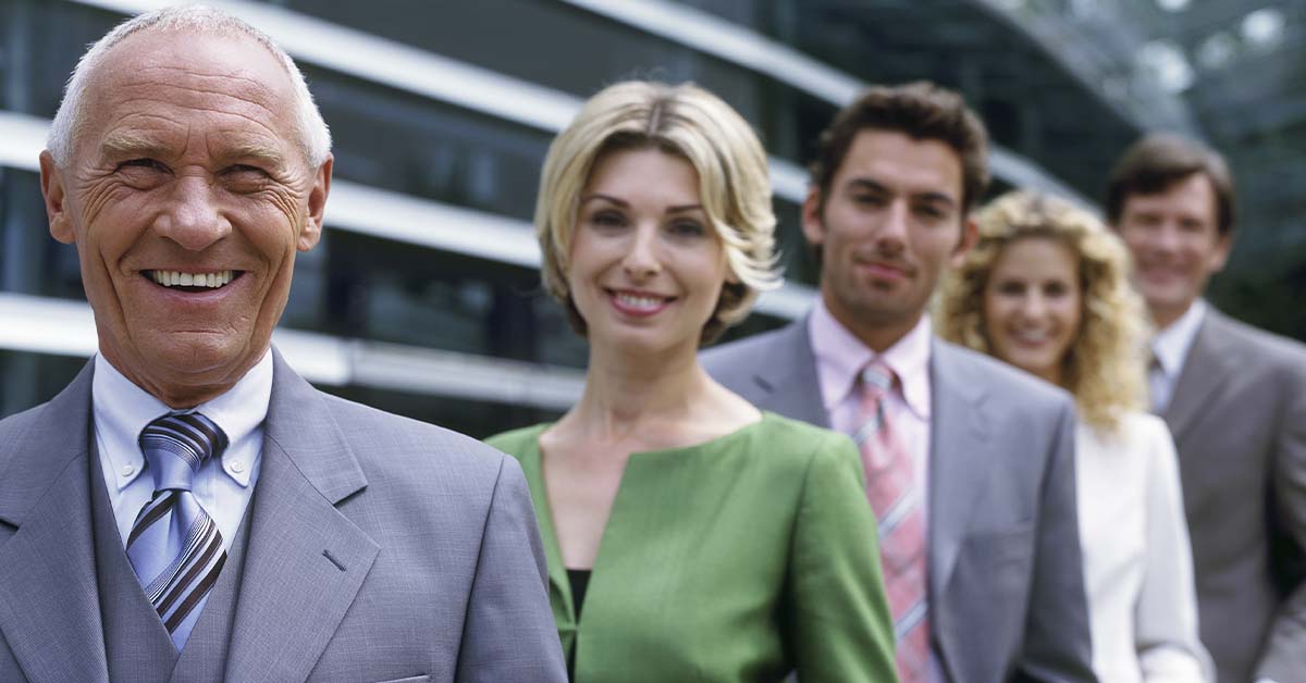 A smiling senior male executive in a grey suit standing in front of a line of blurred colleagues outside a glass building, featured image for the article Therapy for Executives Between Roles.