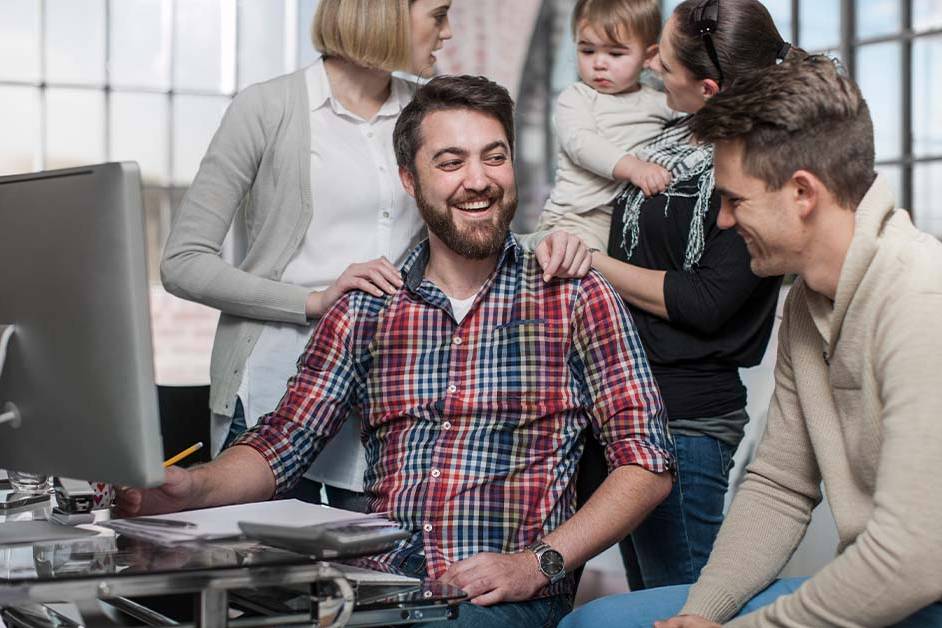 A relaxed group of casually dressed adults, one holding a toddler, gather around a desk with a computer in a bright loft-style office, smiling and talking together.