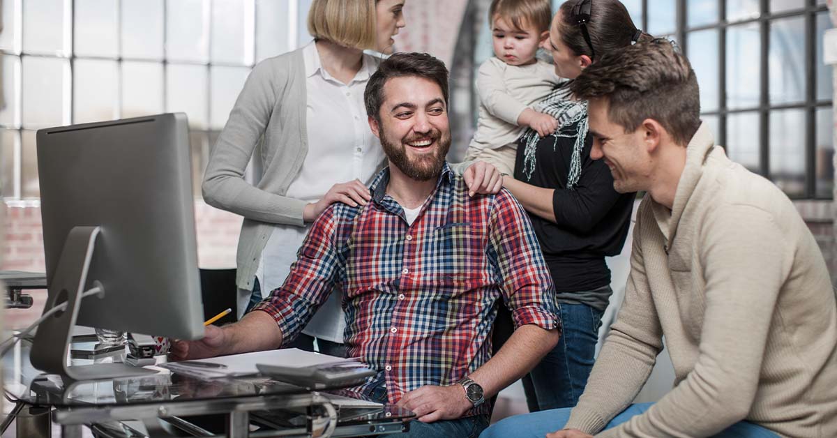 A relaxed group of casually dressed adults, one holding a toddler, gather around a desk with a computer in a bright loft-style office, smiling and talking together.