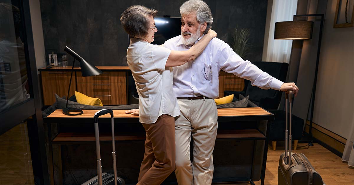 An older couple embracing warmly next to their rolling luggage in a modern living space, featured image for the article Therapy for High-Achievers Returning From Leave.