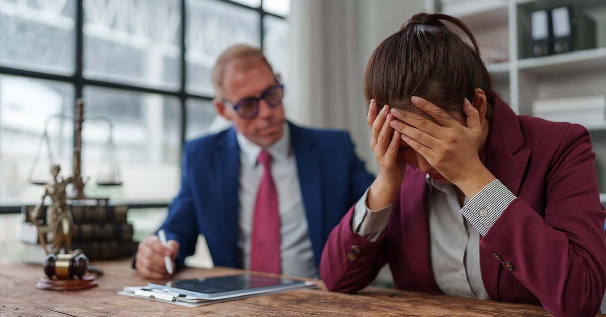 A distressed female lawyer covering her face with her hands at a desk next to a gavel and scales of justice, featured image for the article Therapy for High-Functioning Depression in Lawyers.