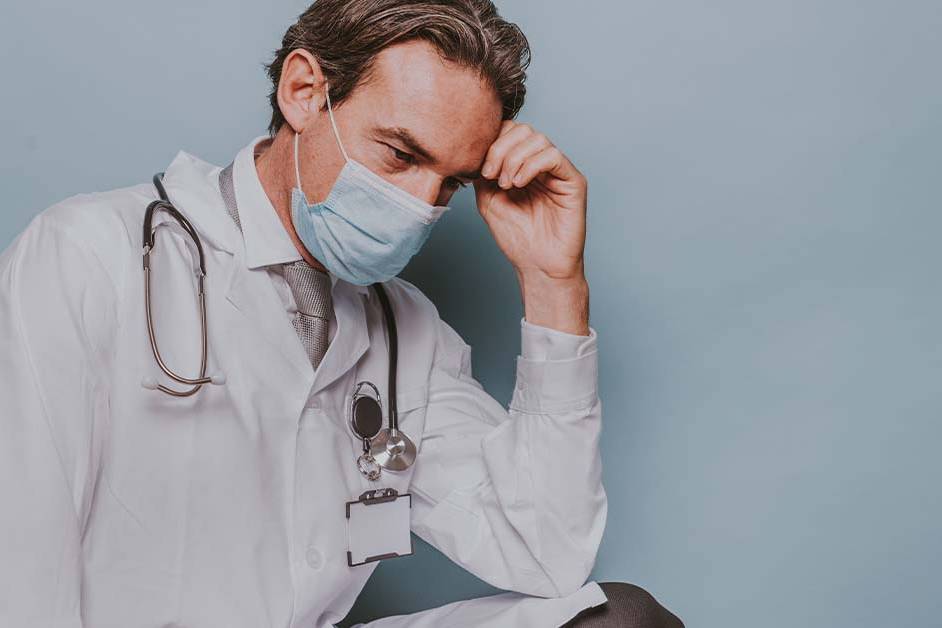 An exhausted male physician in a white lab coat and surgical mask resting his head in his hand, featured image for the article Therapy for High-Functioning Depression in Physicians.