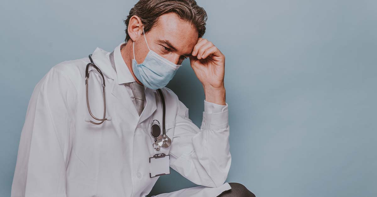 An exhausted male physician in a white lab coat and surgical mask resting his head in his hand, featured image for the article Therapy for High-Functioning Depression in Physicians.