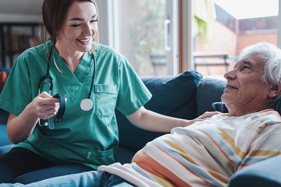 A smiling female nurse in green scrubs sitting next to an elderly man resting on a couch, featured image for the article Therapy for Hospice and Palliative Care Nurses.
