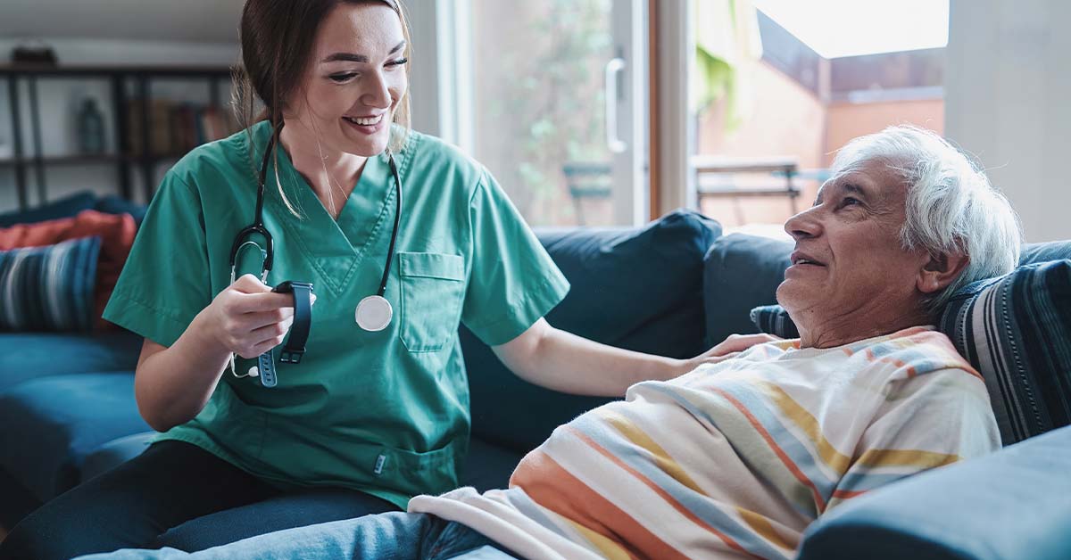 A smiling female nurse in green scrubs sitting next to an elderly man resting on a couch, featured image for the article Therapy for Hospice and Palliative Care Nurses.