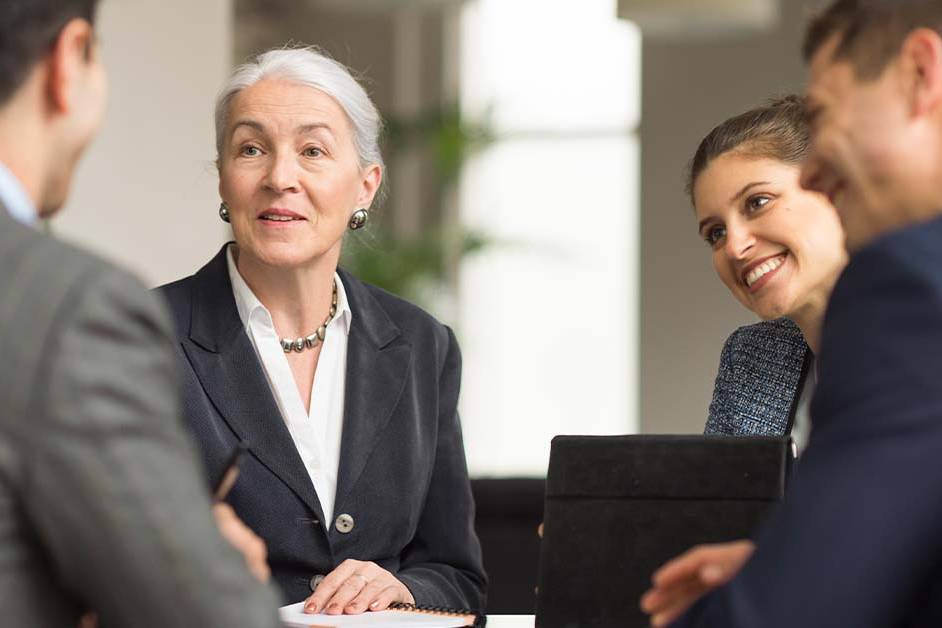 A composed senior businesswoman with grey hair in a dark suit sits across a conference table from colleagues in a meeting, with a younger woman smiling beside her in a bright modern office.