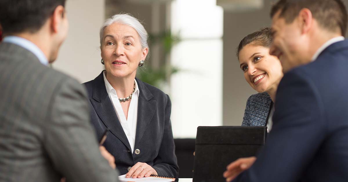 A composed senior businesswoman with grey hair in a dark suit sits across a conference table from colleagues in a meeting, with a younger woman smiling beside her in a bright modern office.