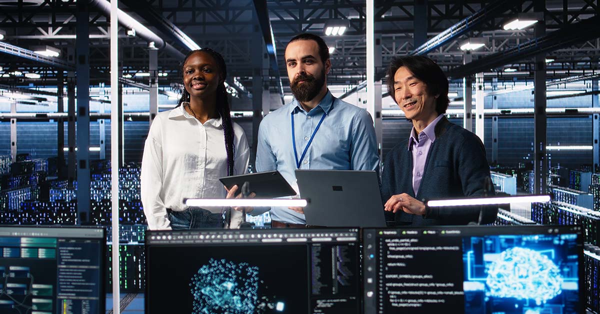 Three diverse tech professionals holding laptops stand in a large data center with rows of servers behind them and monitors showing code and AI brain visualizations in front, representing machine learning engineering.