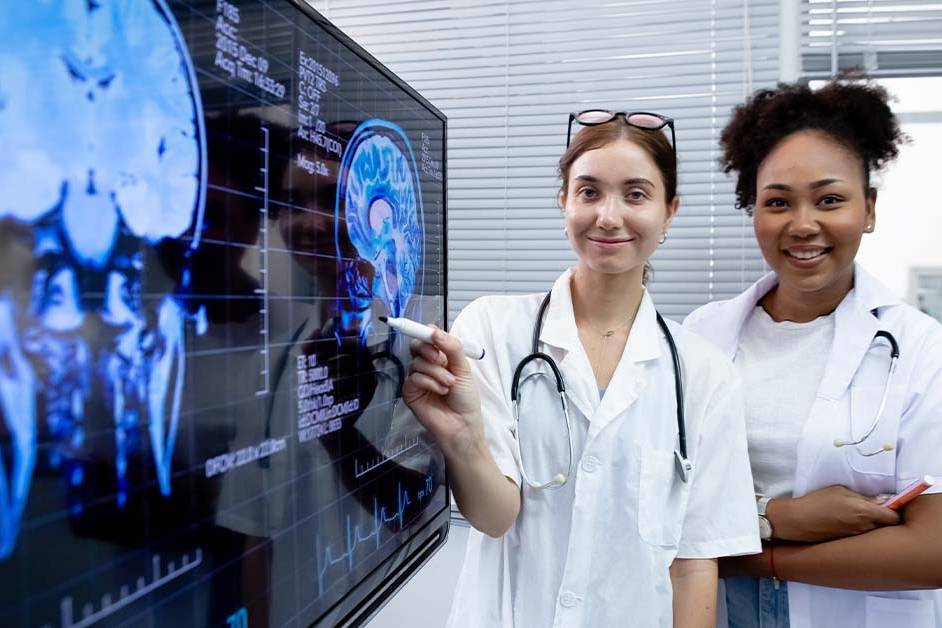 Two smiling female physicians in white coats with stethoscopes stand beside a large display showing brain MRI and CT scans, representing neurosurgical and neurological practice.