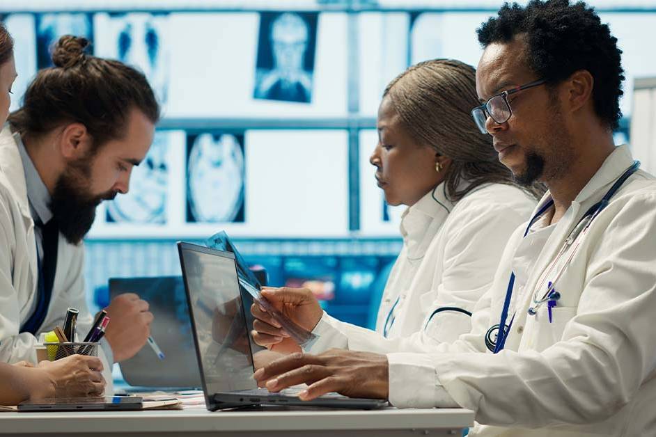 A diverse team of four physicians in white coats sit together reviewing an X-ray film and laptop, with medical scans displayed on screens behind them, representing oncology case review.