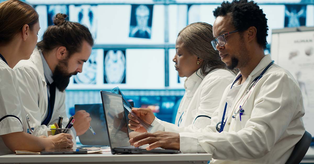 A diverse team of four physicians in white coats sit together reviewing an X-ray film and laptop, with medical scans displayed on screens behind them, representing oncology case review.