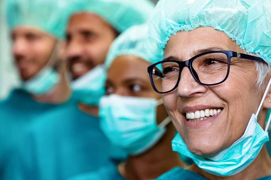 A smiling senior female surgeon in teal scrubs, surgical cap, and glasses stands in the foreground with her mask lowered, alongside a diverse surgical team in caps and masks behind her.