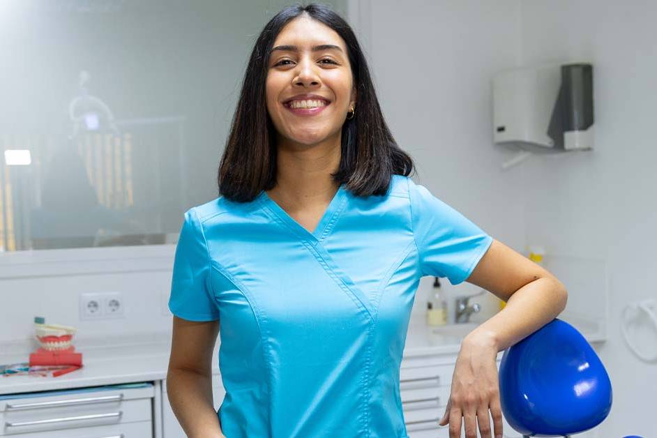 A smiling young female dental professional in blue scrubs stands confidently in a modern dental office, resting one hand on a blue dental chair with a teeth model and equipment behind her.