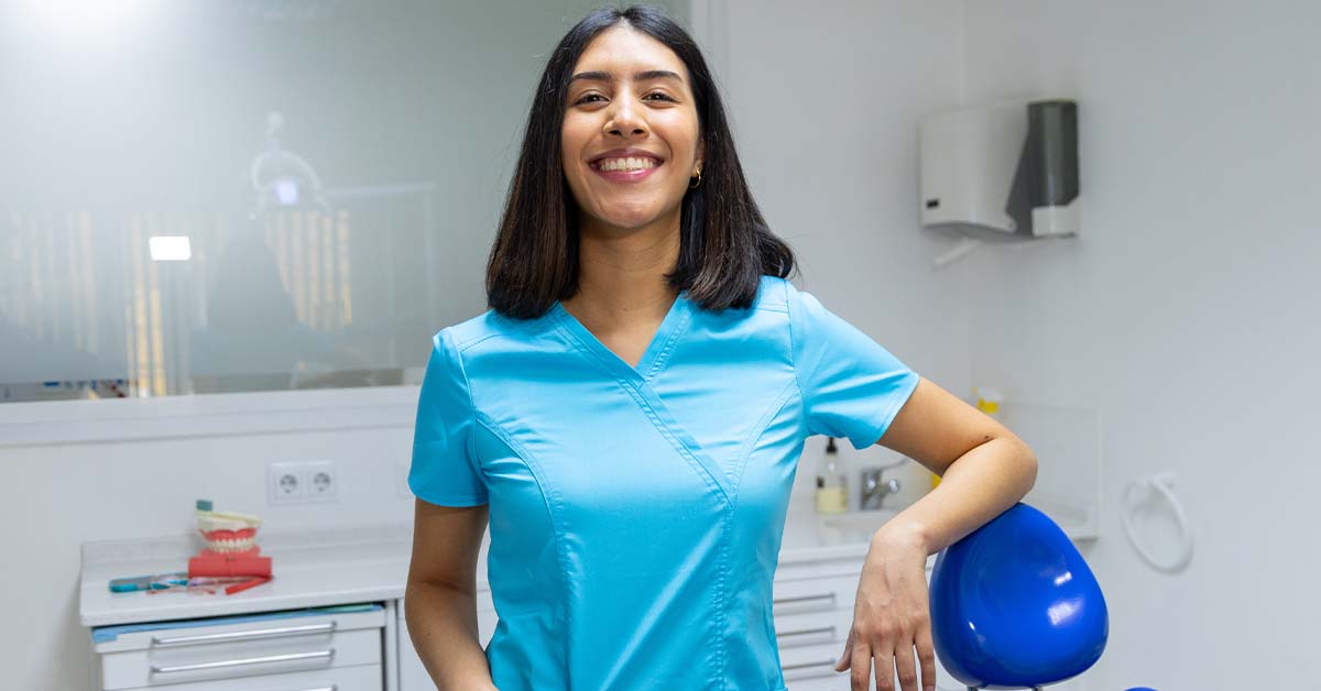 A smiling young female dental professional in blue scrubs stands confidently in a modern dental office, resting one hand on a blue dental chair with a teeth model and equipment behind her.