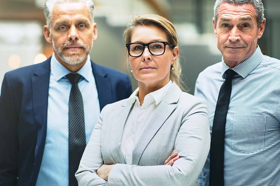 Three serious senior business executives, a woman in glasses flanked by two men in suits, stand with arms crossed looking confidently at the camera in a modern office.