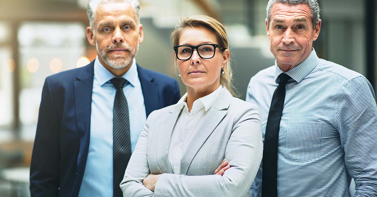 Three serious senior business executives, a woman in glasses flanked by two men in suits, stand with arms crossed looking confidently at the camera in a modern office.
