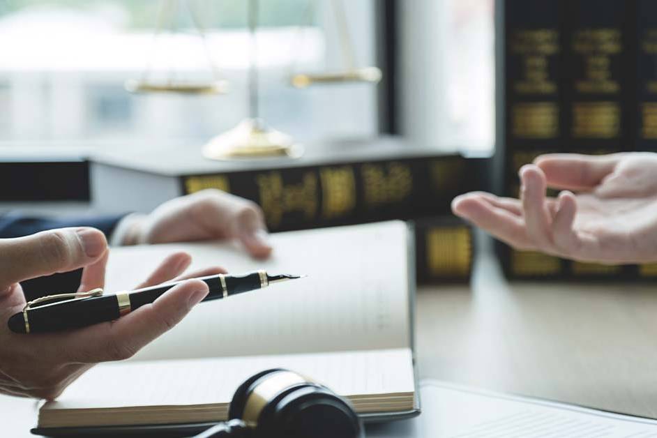 Two people's hands gesture in conversation across a desk holding a fountain pen and open notebook, with a gavel, scales of justice, and shelves of law books in the background.