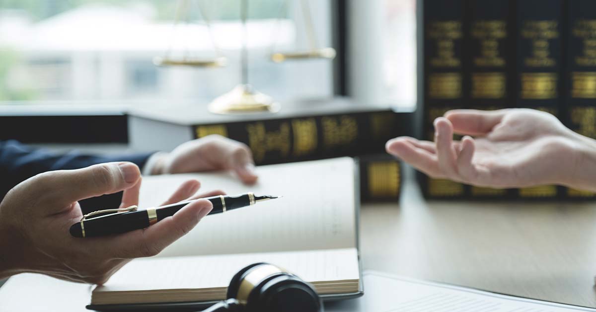 Two people's hands gesture in conversation across a desk holding a fountain pen and open notebook, with a gavel, scales of justice, and shelves of law books in the background.