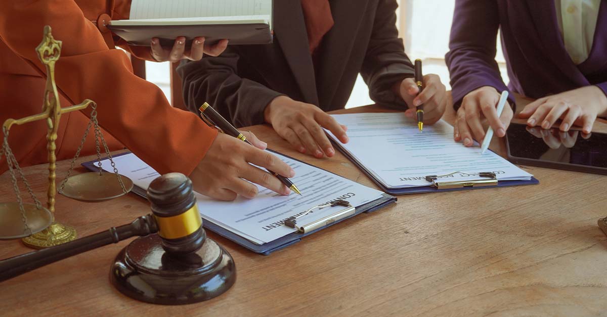 Several attorneys in suits review and sign contract agreement documents at a wooden table, with a brass scales of justice and a gavel in the foreground, representing legal practice.
