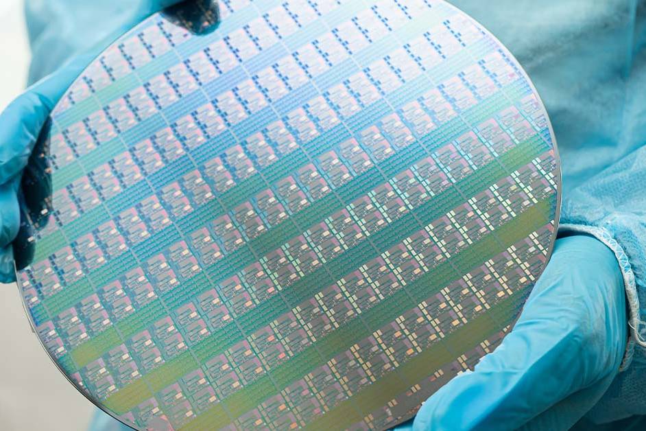 Gloved hands in a blue cleanroom suit hold an iridescent silicon wafer patterned with hundreds of microchips, representing semiconductor manufacturing.