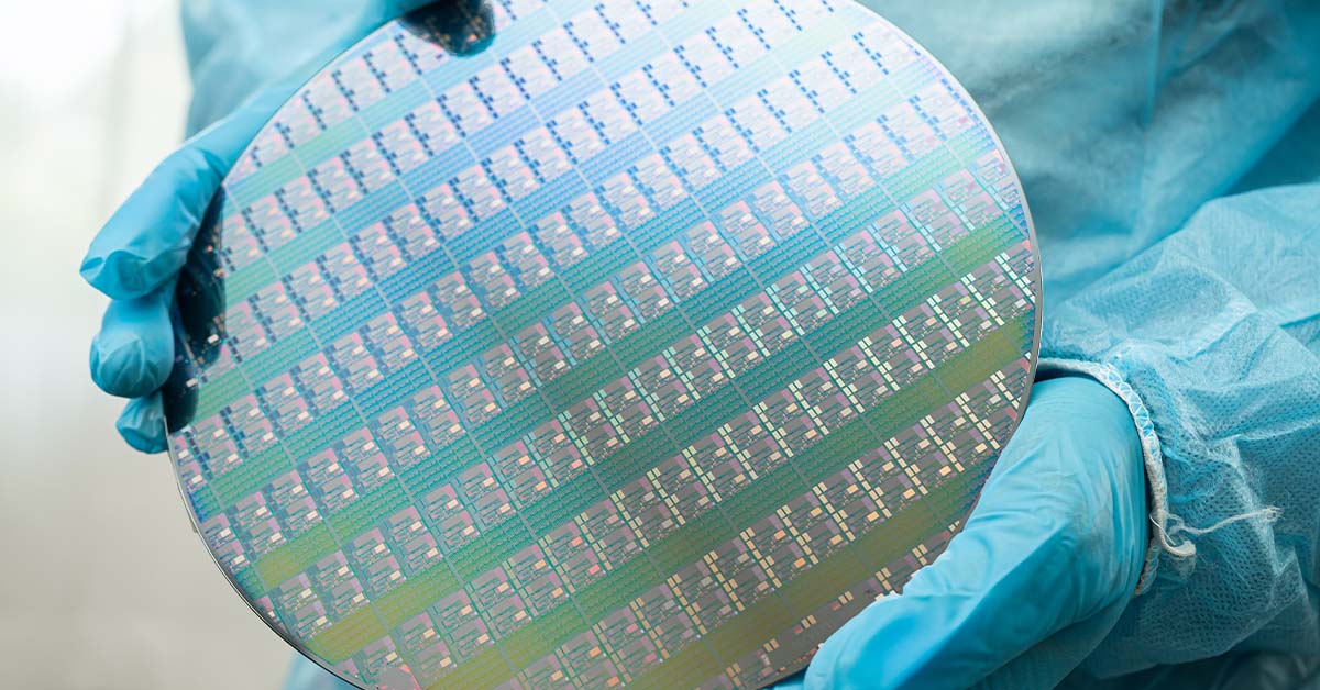 Gloved hands in a blue cleanroom suit hold an iridescent silicon wafer patterned with hundreds of microchips, representing semiconductor manufacturing.